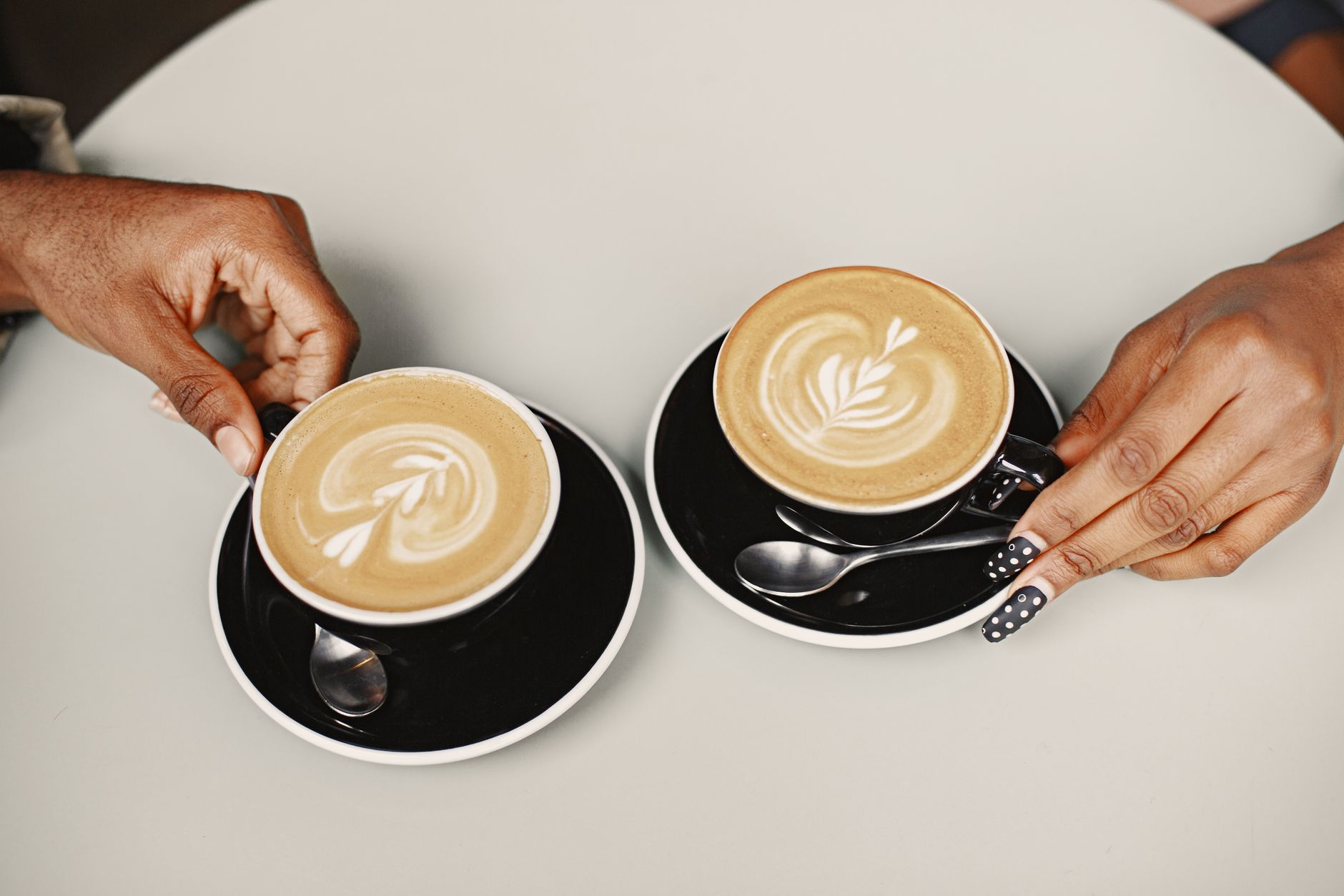 To hænder holder hver sin kop kaffe med latteart på et hvidt bord